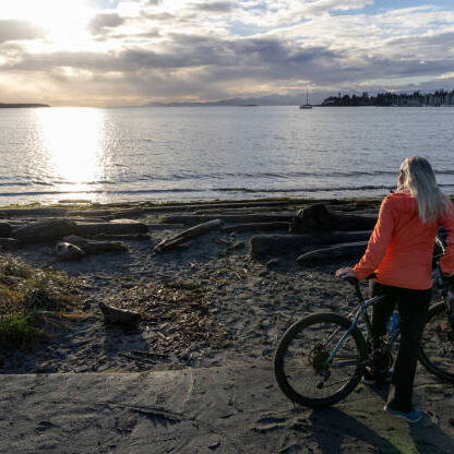 eldery bike Mature woman bicycles along beach at sunrise, calm bay and lofty clouds distant, Victoria, B.C.